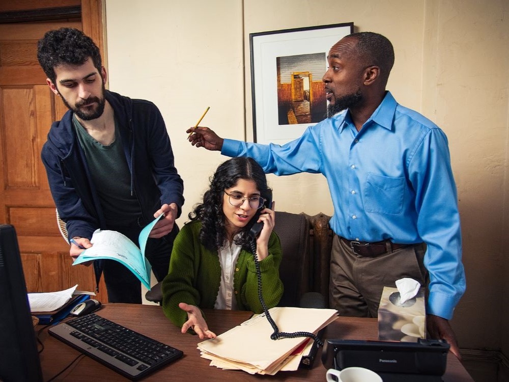 Three people surround a desk. One is flipping through a file folder, one is seated at the desk talking on a phone, and one is gesturing with a pencil in hand.