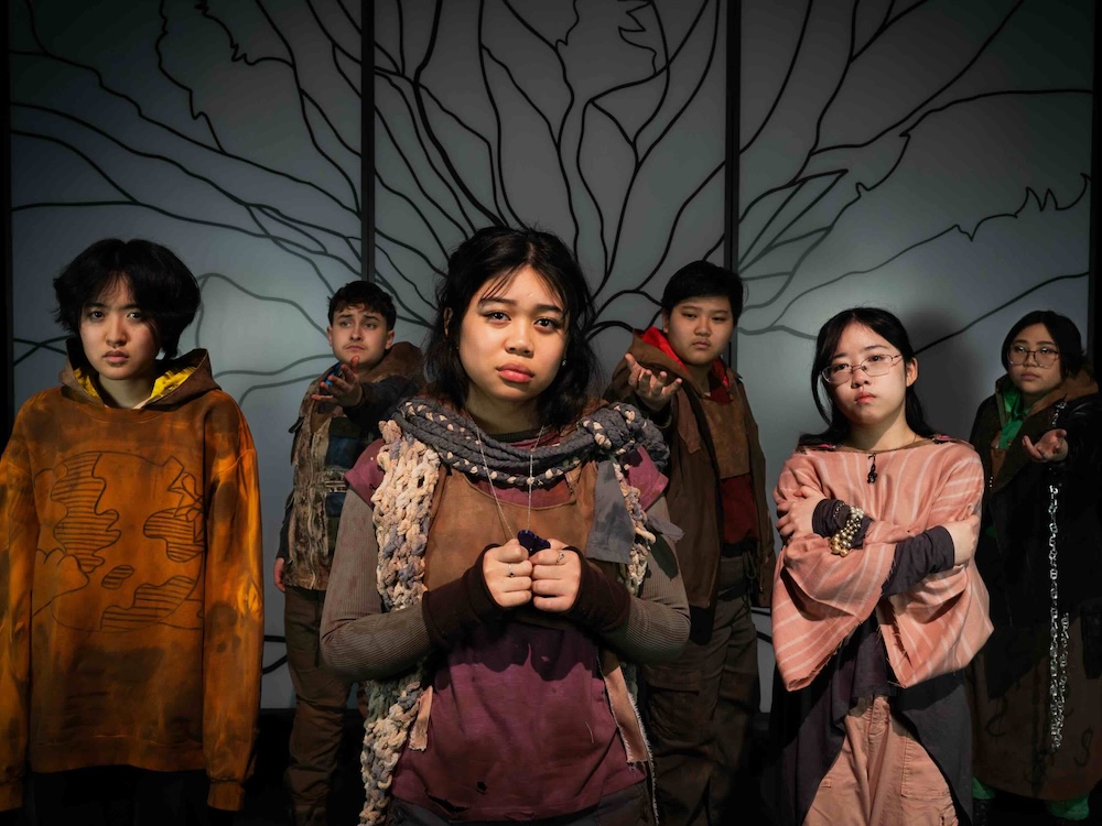 Seven young adults stand together in costume onstage. They are wearing oversized textured clothing in neutral tones. They have dark hair and are standing in front of a grey screen featuring a black illustration of a deciduous tree with many branches.
