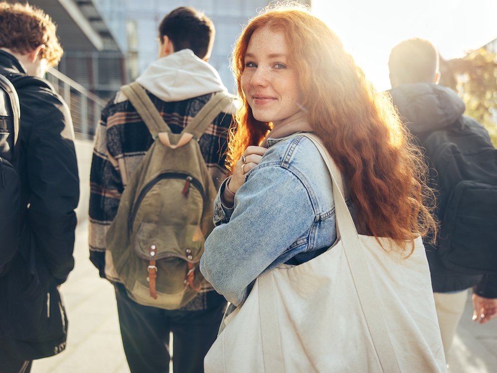 A young woman with long red hair looks backward to smile at the camera. A group of youth wearing backpacks walk ahead of her in the background.
