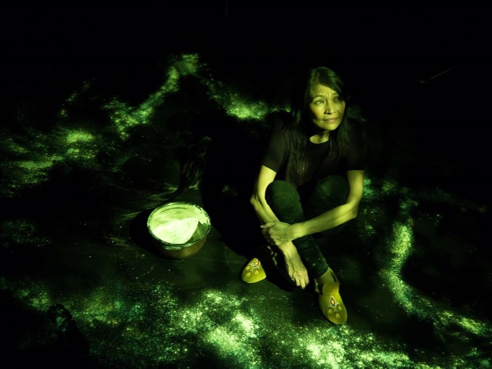 Michelle sits cross-legged onstage wearing black jeans and a black shirt, washed in green theatre light. To her left is a bowl of dough, and flour dust encircles her.