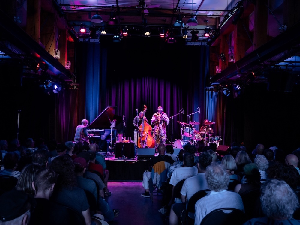 A four-person jazz band, featuring a saxophonist, stand-up bass player, drummer and pianist, play onstage under purple lighting to a full audience.
