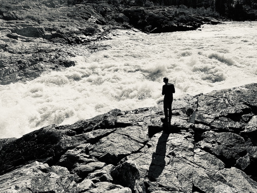 Black and white photograph of a person standing on the rocks by the edge of a whitewater river.