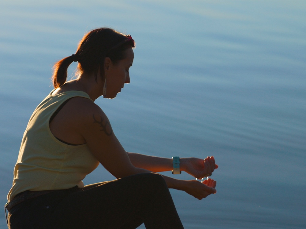 Director Lyana Patrick sits at the edge of a lake, cupping some of the water in her hand. The water is still and there is sunlight in her hair.