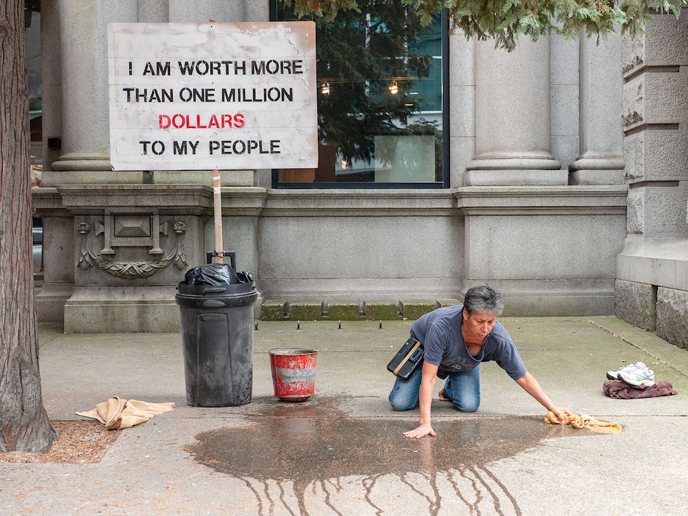 Dressed in jeans and a blue T-shirt, artist Rebecca Belmore takes a wet cloth to a city sidewalk. Behind her is a sign, held up by a black garbage can, that reads ‘I am worth more than one million dollars to my people,’ with the word ‘dollars’ highlighted in red.