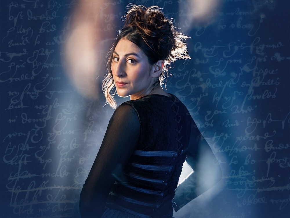 A woman with medium-light skin with dark hair in an updo stands with her back to the camera and her head turned to look into the camera. The image is superimposed on a blue background covered with illegible handwriting.