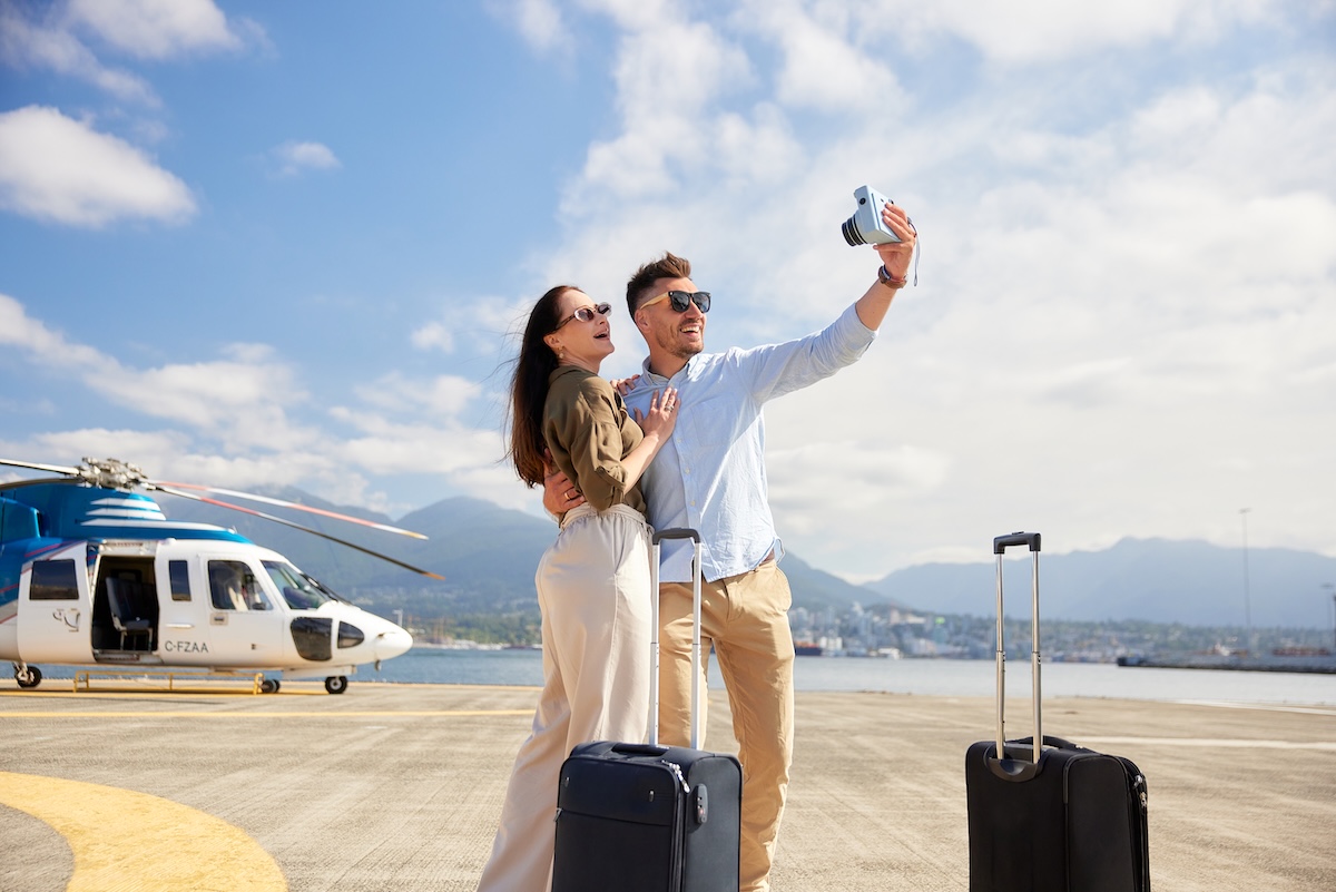A couple with carry-on suitcases near them on a helicopter pad takes a selfie with a helicopter, water and mountains in the background.