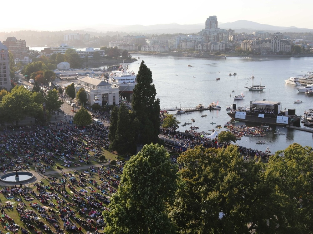 Aerial view of crowds of people facing a symphony playing on a barge on a city harbour.