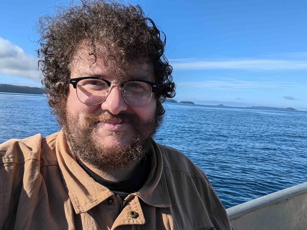 Tanner Zurkoski has brown curly hair and glasses. He is wearing a brown button-down shirt and standing near a railing by the water. The sky behind him is bright blue.