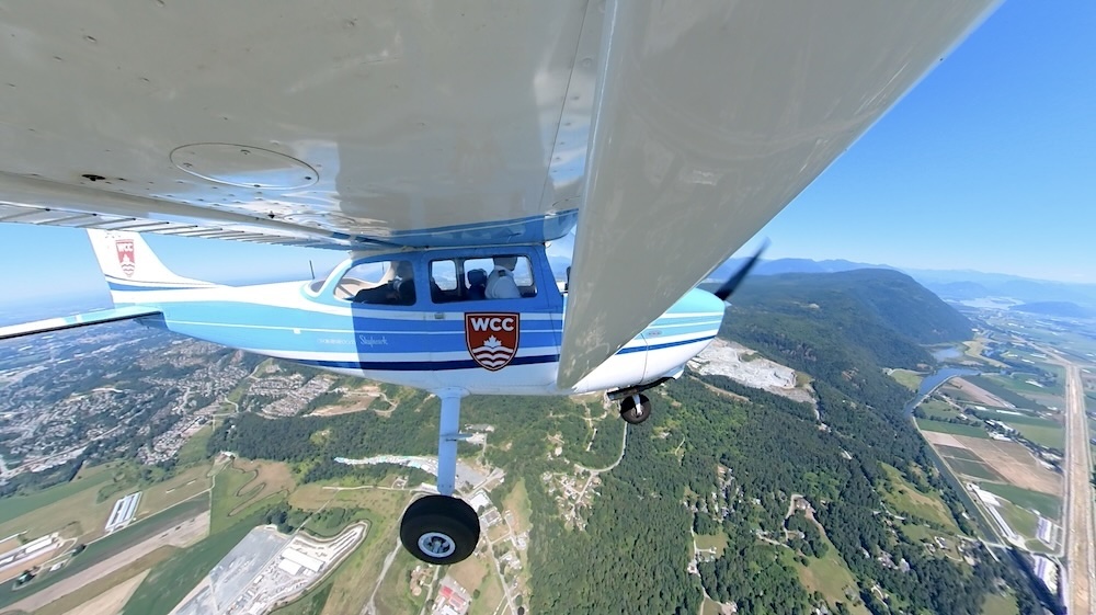 A small plane with a WCC logo in flight over green hills and farmland.