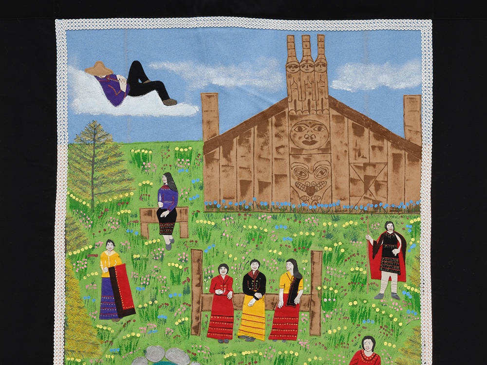 A brightly coloured blanket, with a thick black border, depicting seven figures relaxing in a Haida Gwaii landscape with a traditional big house in the background.