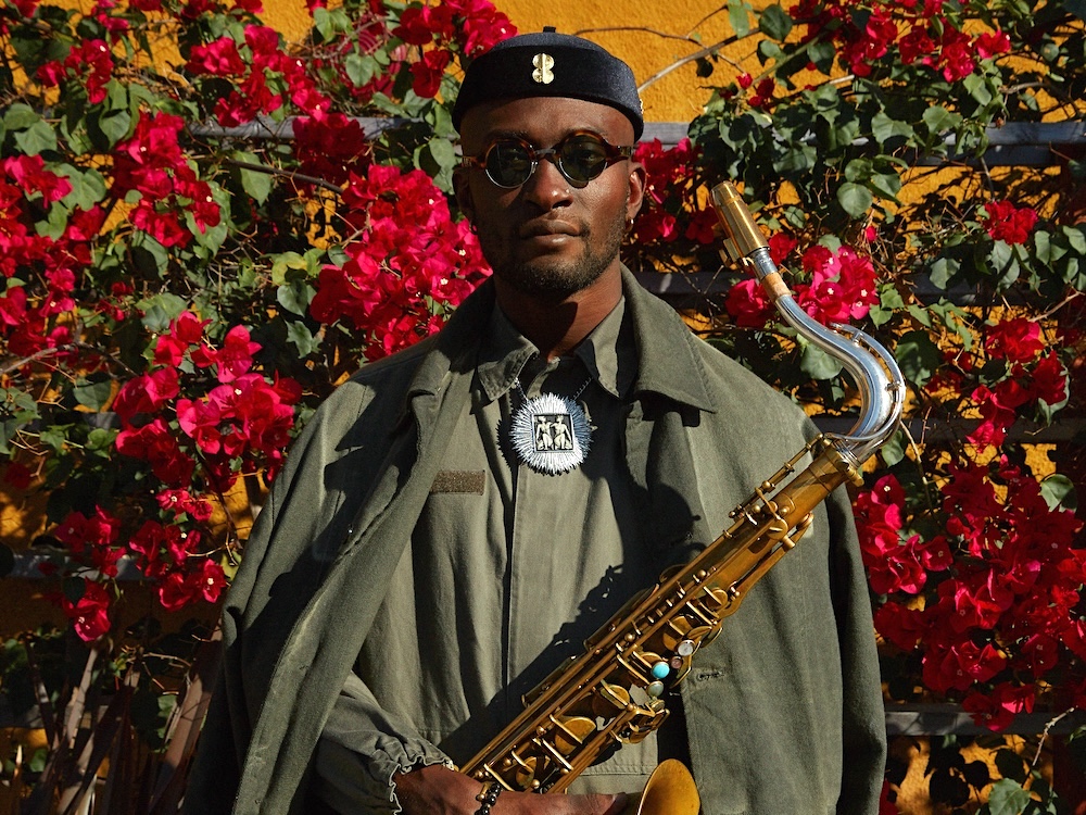 Isaiah Collier stands in front of a flower-covered fence holding a saxophone. He looks directly at the camera and wears an army-green shirt and jacket, a black cap and black sunglasses.