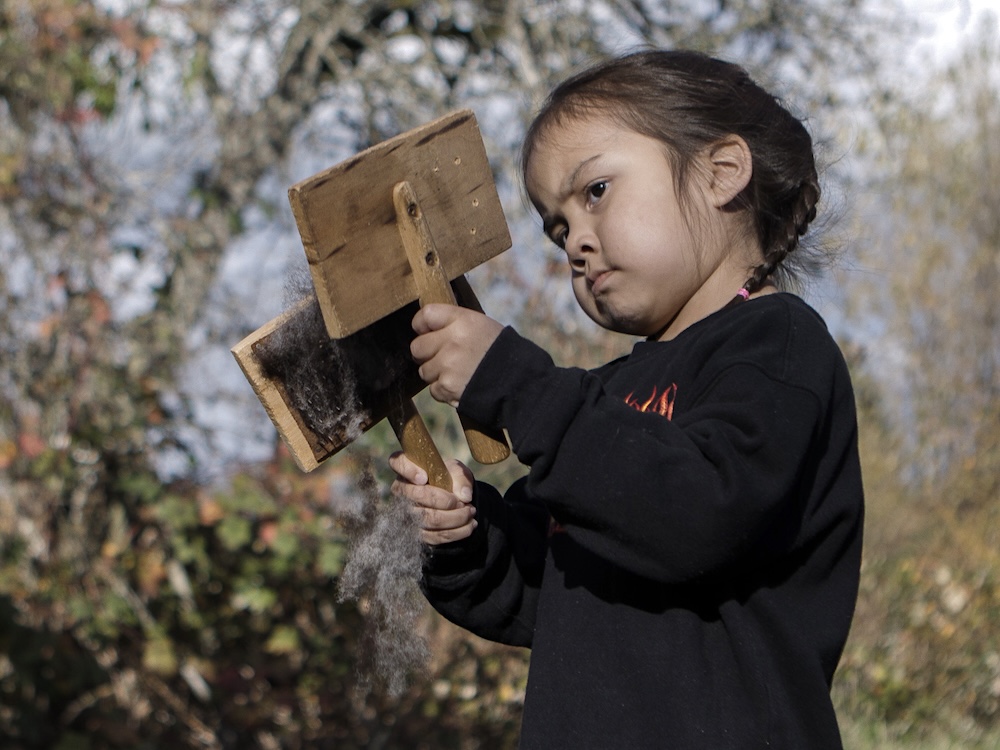 A child with long dark hair pulled back holds two wood combers full of wool in each hand. She stands outdoors against a forested backdrop. 
