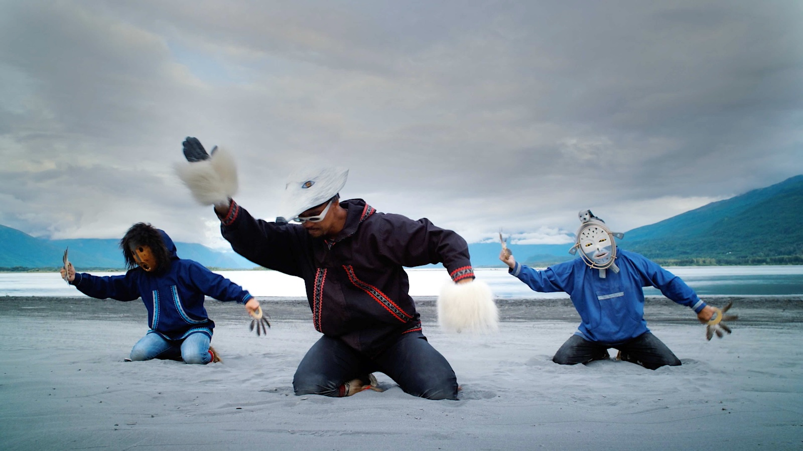 Three people kneel in snow, mid-dance. They are performing as part of a musical group, and the two people on the left and right side are wearing masks carved by Yup&rsquo;ik artist Drew Michael. Behind them is water, mountains and an overcast sky.
