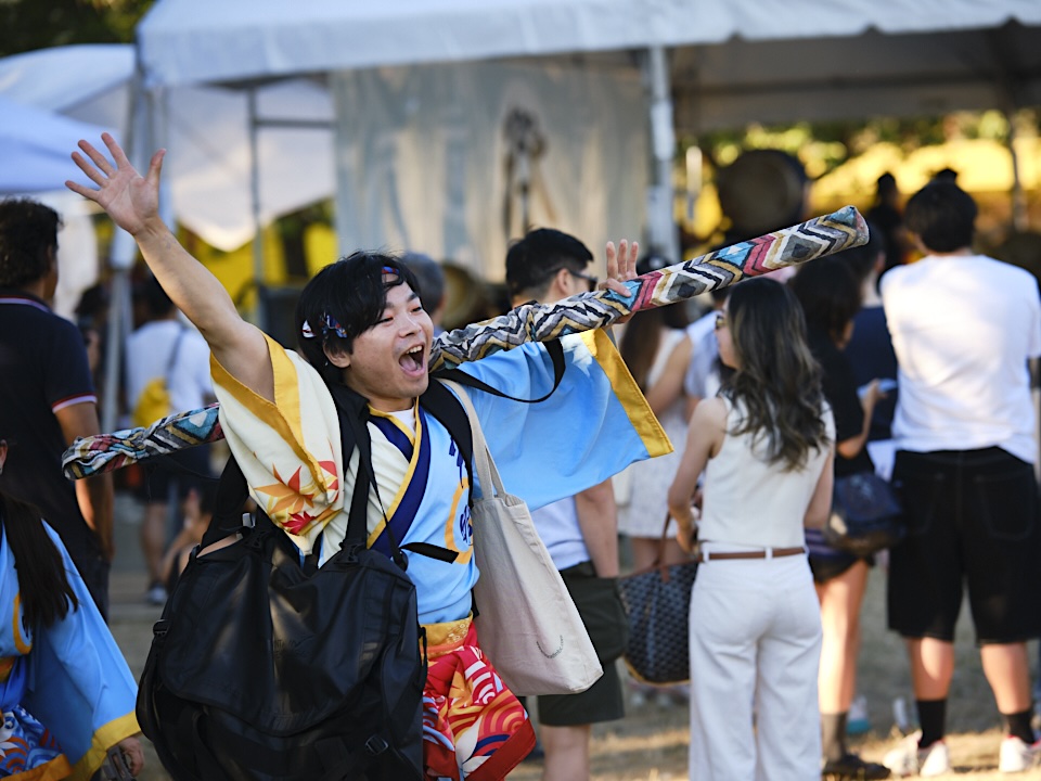 A person in traditional Japanese dress holds their arms in the air in greeting and celebration. They&rsquo;re smiling. Behind them are people in a festival environment among white tents in a park.