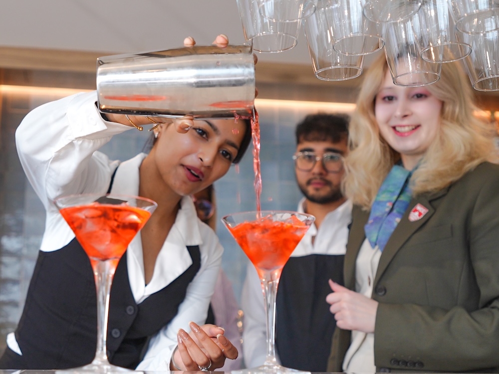 A bartender pours two orange-coloured cocktails out of a shaker. 