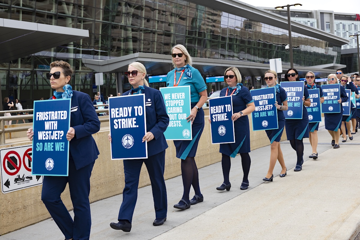 WestJet attendants walk in a line in front of an airport, holding signs that say &lsquo;Frustrated with WestJet? So are we!&rsquo; and &lsquo;Ready to strike.&rsquo;