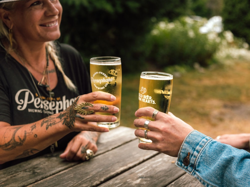 Two people sit across from each other at a wooden table outdoors. They are holding up tall pint glasses from Persephone Brewing Co., as if to say, &lsquo;Cheers.&rsquo;