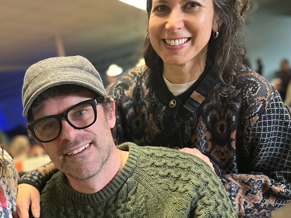 Brian Smith, at left, has short brown hair, a grey tweed cap and glasses. He is seated and wearing a green sweater. Melissa Smith, at right, stands behind him. She has wavy dark hair and is wearing a patterned cardigan.