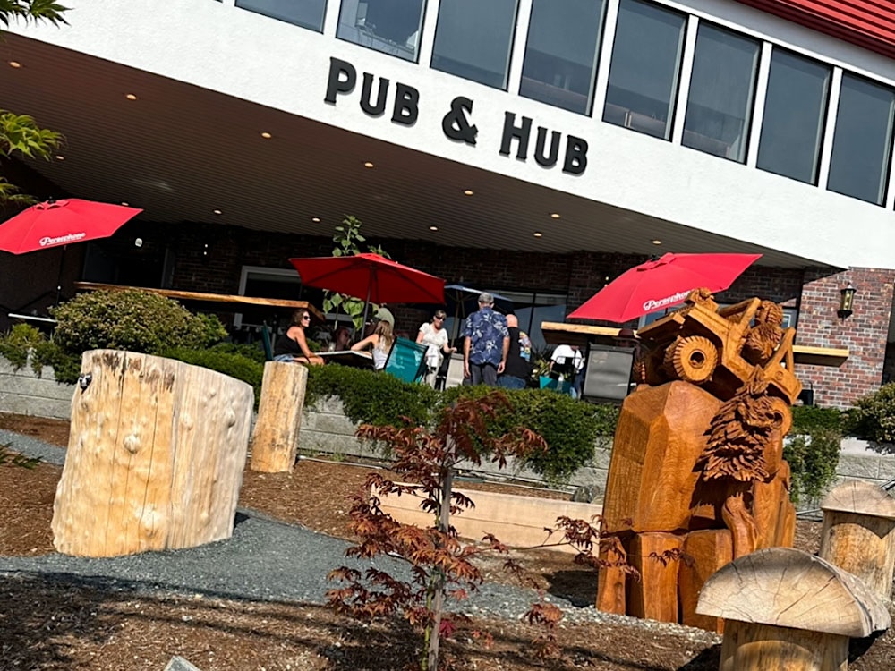 The exterior of a white building with a sign that reads &lsquo;Pub & Hub&rsquo; features a patio on the ground floor with red Persephone Brewing umbrellas.