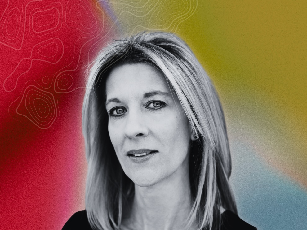 A black and white headshot of a woman with light skin tone and shoulder-length blond hair, on a multicoloured background.