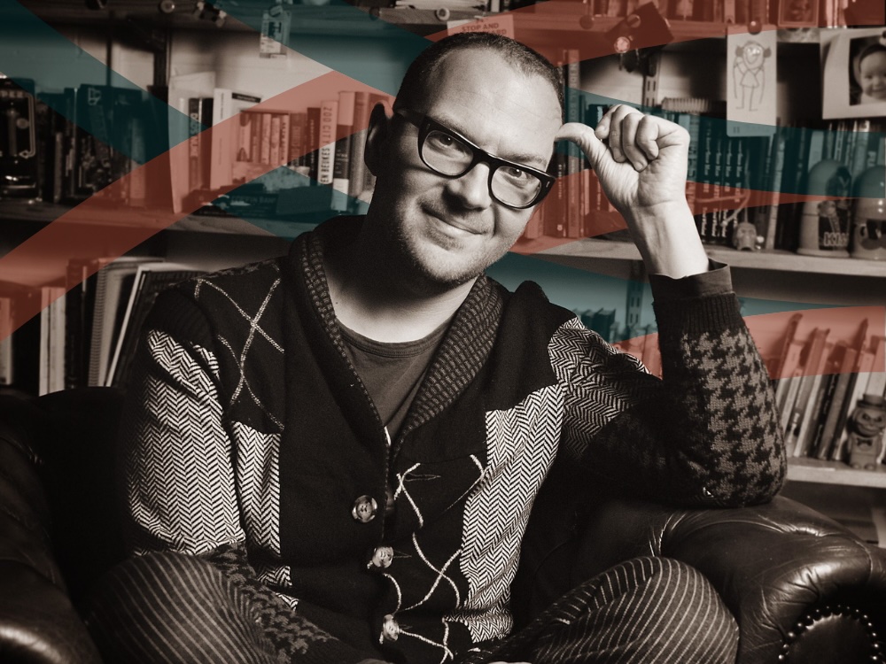 Cory Doctorow smiles for the camera. He sits in a leather chair and wears a multi-patterned cardigan.