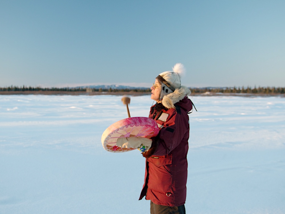 A person stands holding a drum in an icy field against a blue sky. 