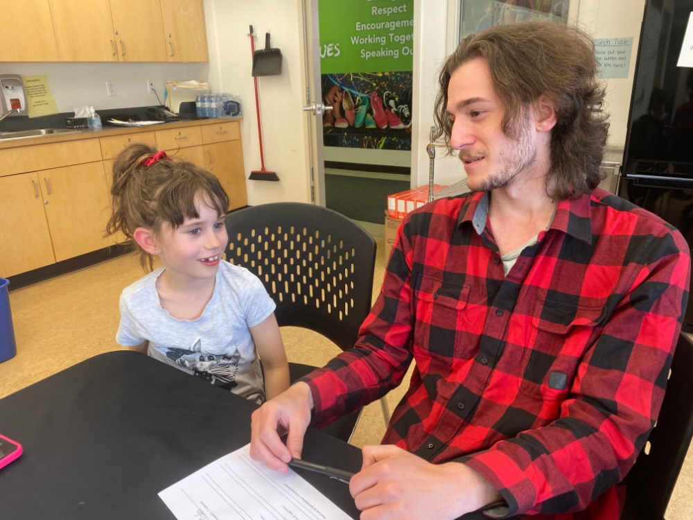 A young man in a red plaid shirt, at right, sits next to a young girl at a table in an indoor space. They are both smiling, mid-speech.