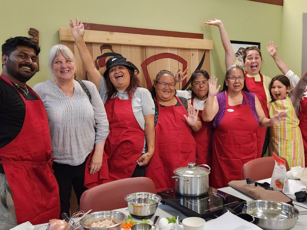 Several people in red aprons pose for a group photo in a community kitchen space. Cooking equipment and ingredients are on the white table in front of them.