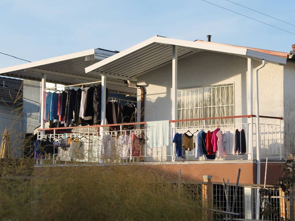 Laundry hangs on the railings and on racks across the back second-storey balcony of a Vancouver Special home in East Vancouver. The exterior of the home is white and there is a lush green bush in the foreground. The sky is blue. 