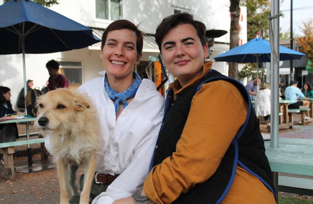 Lizzy Karp, left, and Emma Devin, right, are seated outside at a picnic table with Lizzy&rsquo;s scruffy terrier, who Karp is holding to the left of the frame. Karp has short dark hair, is wearing a white button-down shirt with a blue scarf and smiling. Devin has short dark hair and is wearing a black vest over a pumpkin-coloured button-down shirt. Behind them is a white building and other people at picnic tables.