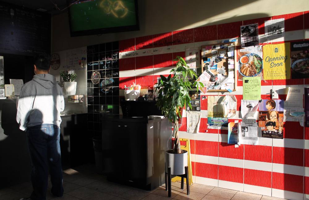Three stacked photos from the Renfrew-Collingwood neighbourhood in Vancouver depict, from top: a colourful collection of plastic step-stools in front of a storefront; a family of two parents and a toddler walking, shot from the back with restaurant signage that reads &ldquo;Congee Noodle King&rdquo; in the foreground to the left of the frame; the interior of an eatery in which one person in grey stands at the counter to the left of the frame while a poster board with a red and yellow sign that reads &ldquo;Opening soon&rdquo; is lit with sunlight streaming in from the right.
