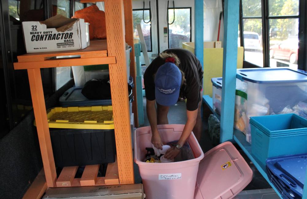 Three stacked photos show the Distro Disco donation hub in action. Top: a spraypainted sign leaning against a grey folding table reads &ldquo;Distro Disco Mobile Store&rdquo; in red, green and blue lettering. Middle: a view of the Distro Disco bus, a repurposed handyDart-style shuttle bus with a large waterproof poster hung across its passenger windows in yellow, orange and green colourways. The sign explains what Distro Disco is about. Bottom: A person in a blue ball cap bends over and reaches into a baby pink Rubbermaid Roughneck container. They are surrounded by a large orange shelf to the left of the frame and a large blue shelf to the right. They are standing in the interior of a repurposed HandyDART van.