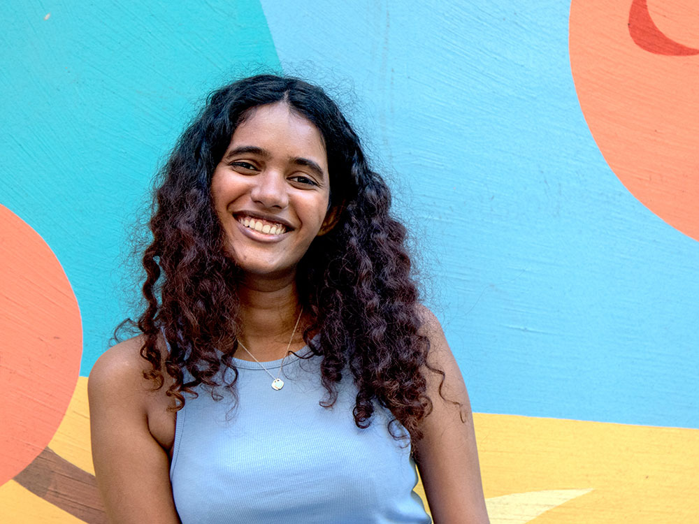 Akhila Menon is looking at the camera and smiling, her head tilted slightly to the left. Behind her are tableside lamps and a cozy restaurant interior.