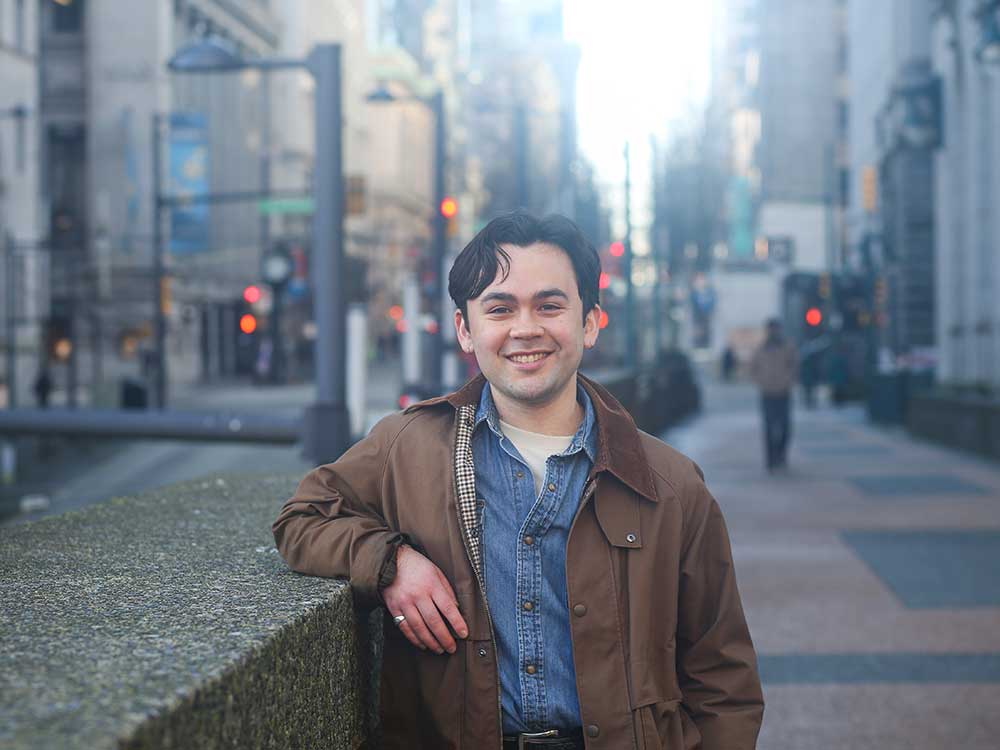 A young man with medium-coloured skin, dark hair and a mushroom cut leans on a stone wall. He is standing on a city street and wearing a blue denim button-up top and an open brown jacket.