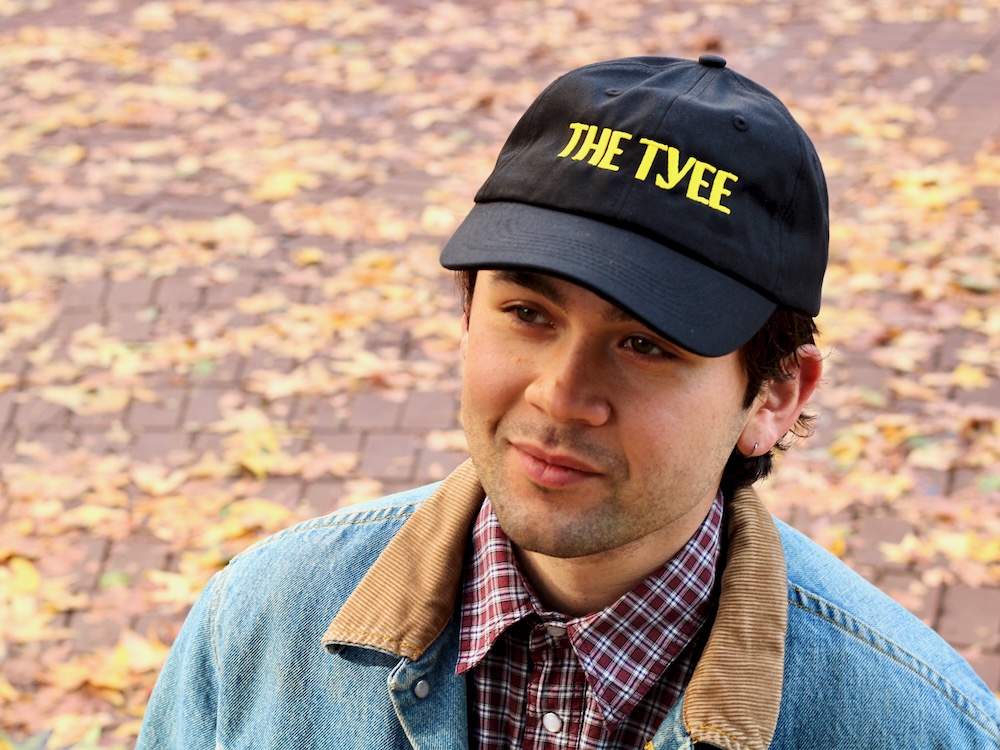 A handsome young man sports a black baseball cap with &lsquo;The Tyee&rsquo; in yellow.