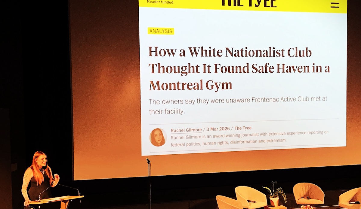 Rachel Gilmore speaks from a lectern at the bottom left corner of the frame. To the right a large screen projects an image of a Tyee article by Gilmore titled &lsquo;How a White Nationalist Club Thought It Found Safe Haven in a Montreal Gym.&rsquo;
