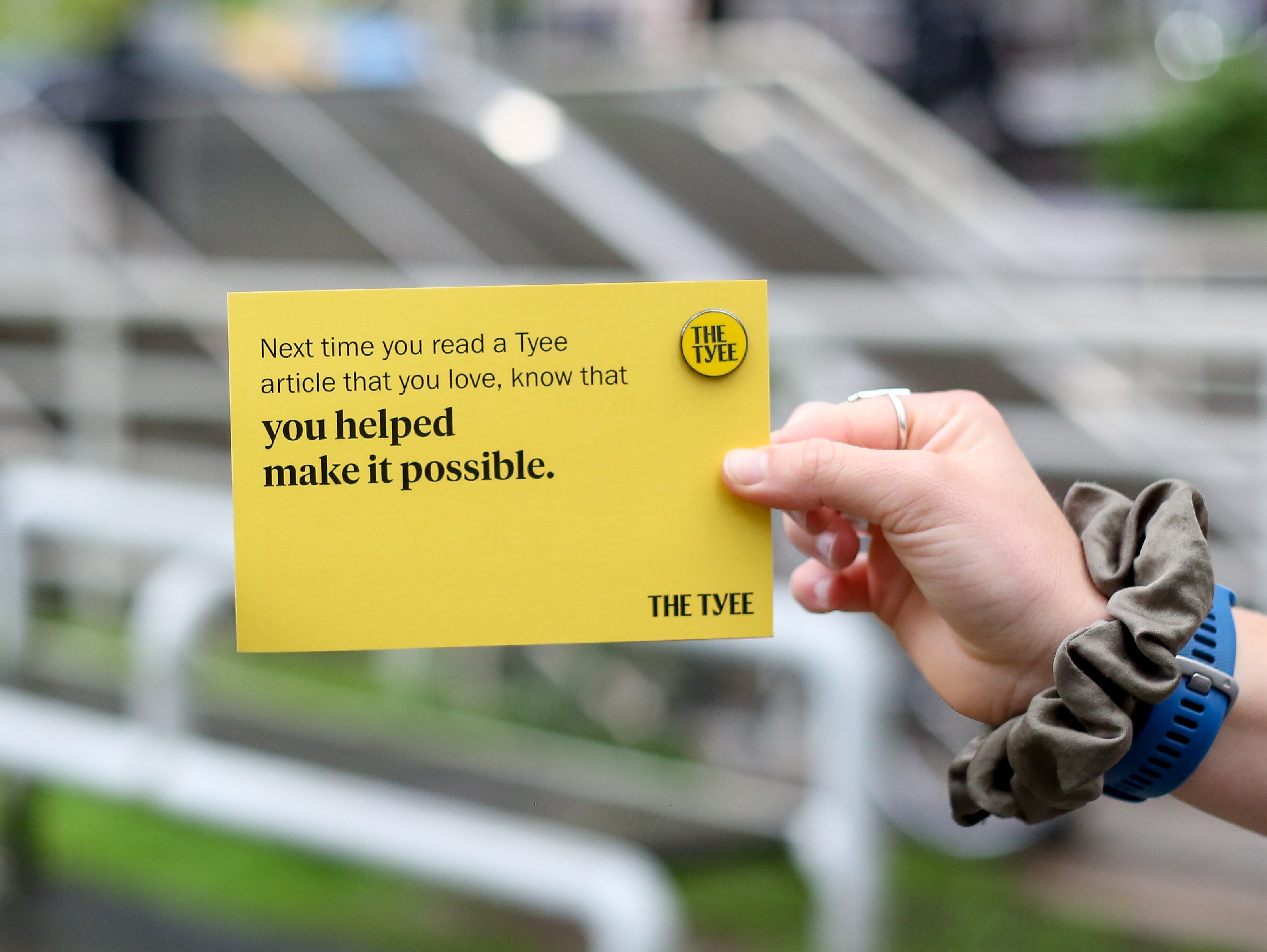 A hand holds a yellow card that reads, &lsquo;Next time you read a Tyee article that you love, know that you helped make it possible,&rsquo; with a Tyee pin attached in one corner