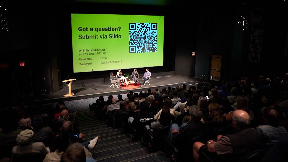 An audience full of people in tiered seats watch four people in discussion onstage.
