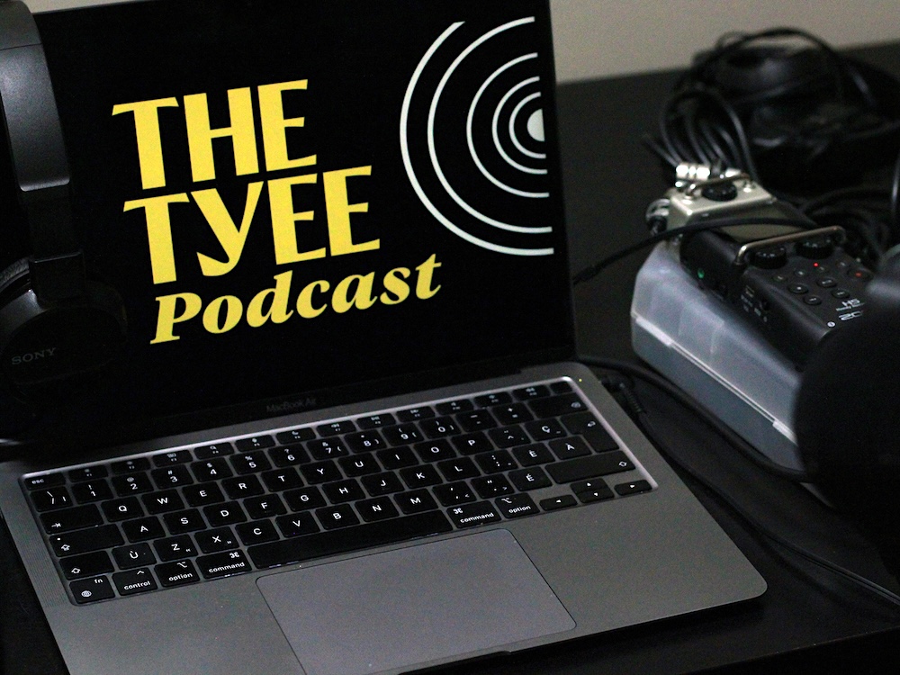 A MacBook Air laptop sits on a black desk next to podcast recording equipment to the right of the frame. On the laptop screen is a black full-screen logo that says &lsquo;The Tyee Podcast&rsquo; in yellow script with a circular white graphic to its right.