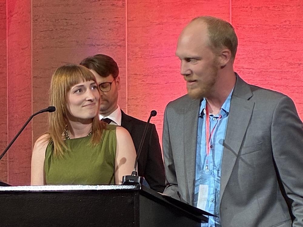 A woman with chestnut hair wearing a green dress stands at a podium beside a man with a receding hairline and trimmed beard.