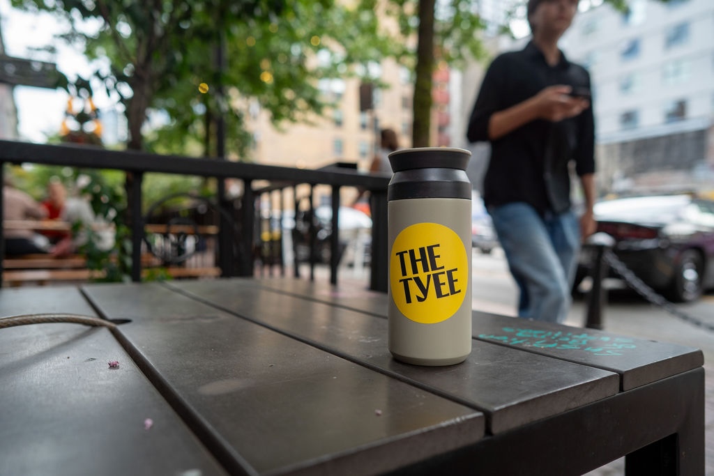 A taupe commuter cup features a circular yellow Tyee sticker on a dark picnic table outdoors. A person in a black shirt and jeans is walking by in the background.