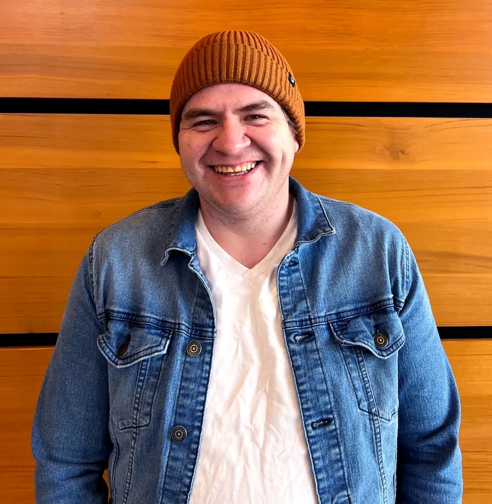 Kevin O'Neill, wearing a brownish-orange toque and a jean jacket over a white T-shirt, smiles directly at the camera.