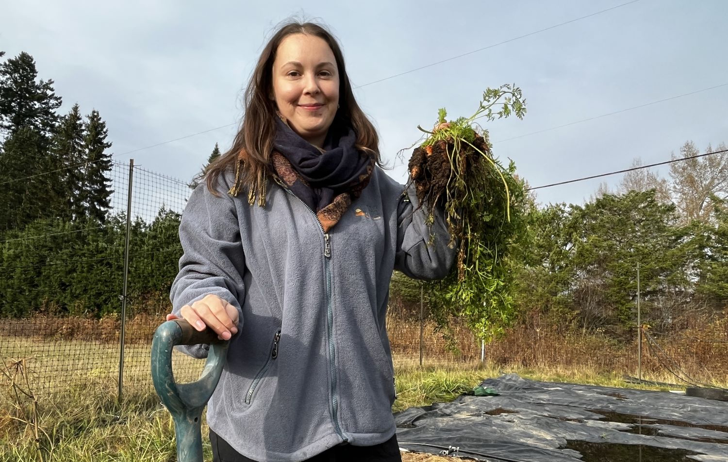 A young Indigenous woman with long dark hair, wearing a grey fleece jacket and scarves, stands in a farm field. Her right hand rests on a shovel; her left holds newly picked carrots.