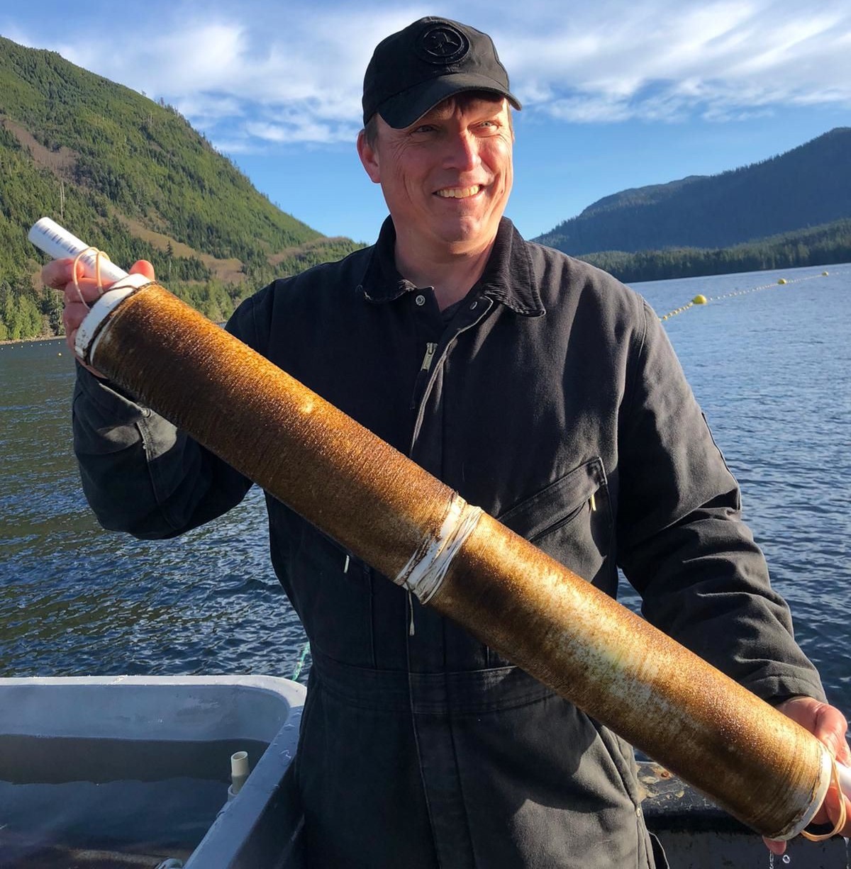 A light-skinned man in a black baseball cap and black coveralls holds a spool covered with brown kelp seed. Water and evergreen covered hills can be seen behind him.