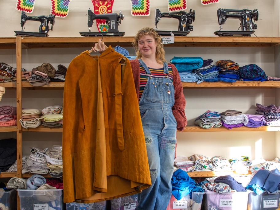 Arora Timberlyn has curly shoulder-length blond hair and light skin. She stands in front of a honey-coloured wooden shelf containing bolts of colourful fabric. A row of vintage black sewing machines lines the top of the shelf under a colourful string of embroidered rainbow bunting. Timberlyn is wearing blue denim overalls over a colourful striped T-shirt and thick pumpkin cardigan. She holds a rust-coloured leather cape on a wooden hanger.