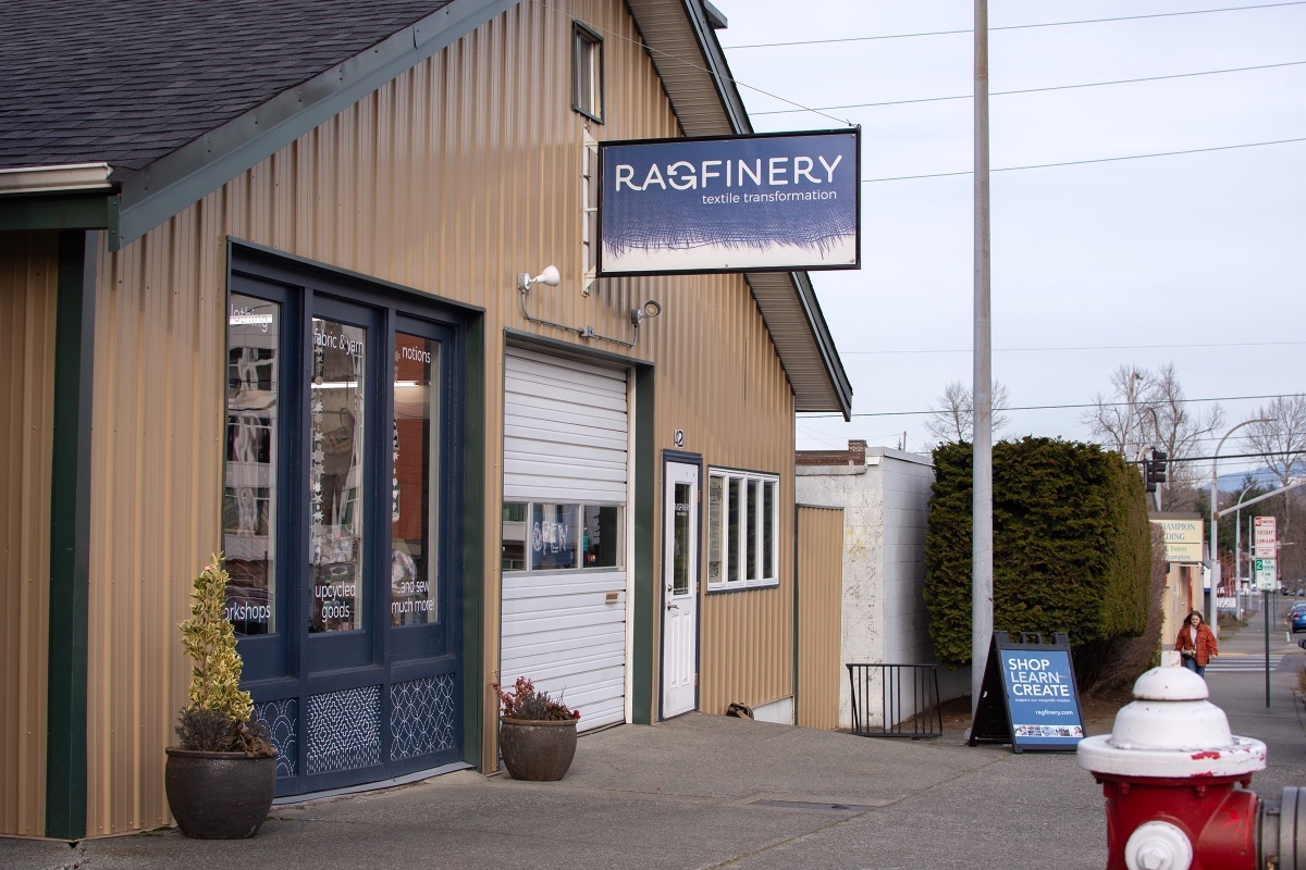 The exterior of the Ragfinery, a two-storey building on a Bellingham street with a peaked roof and beige vinyl siding with dark blue trim and a white roll-up garage door. A blue rectangular sign with the word 'Ragfinery' hangs from the second storey. It&rsquo;s a dry, crisp winter day and the sky is light blue.