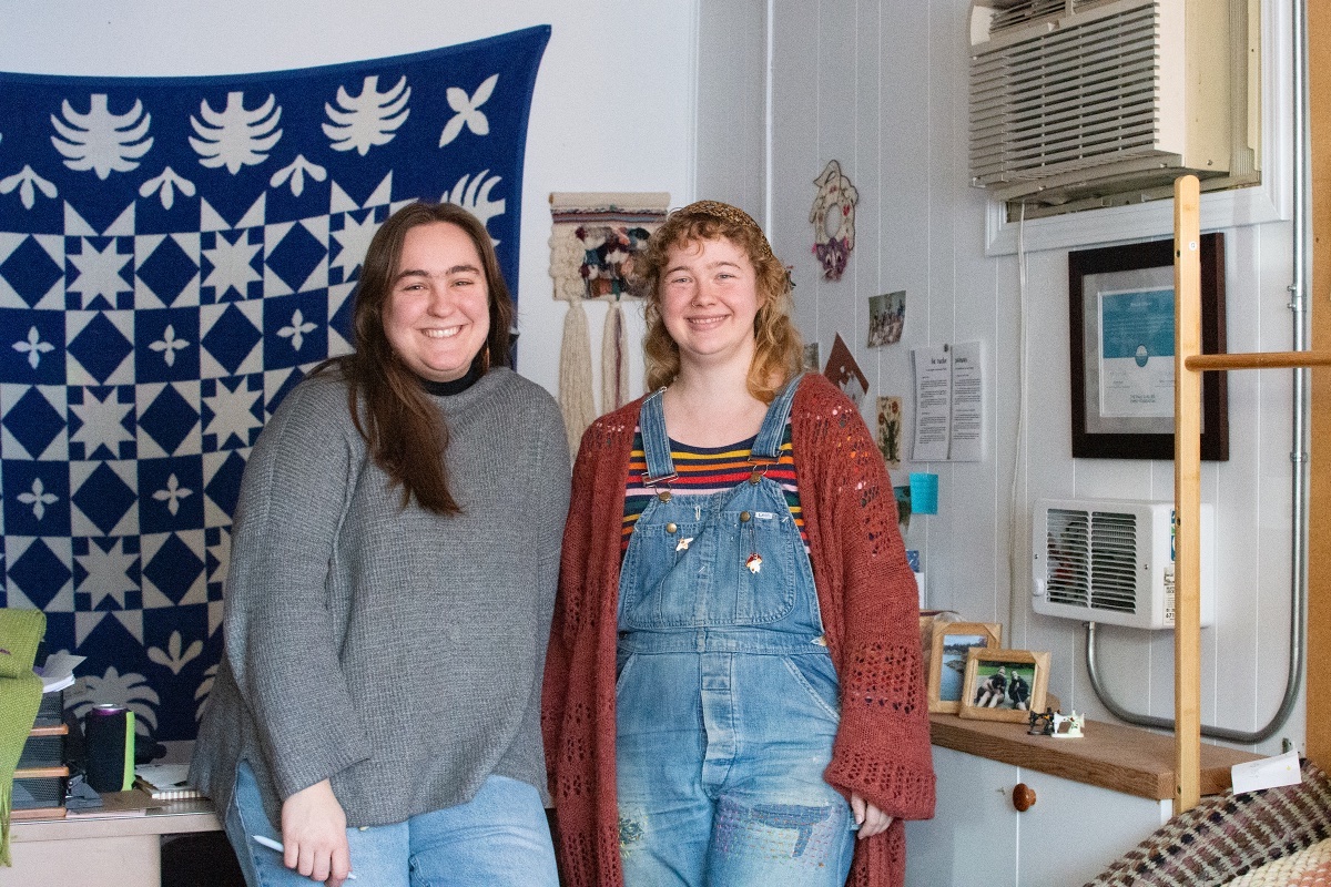 Two people stand and smile brightly at the camera. Rue MaCall, left, has light skin and long brown hair. She is wearing an oversized grey waffle knit sweater and blue jeans. Arora Timberlyn, right, stands beside her. She has curly shoulder-length blond hair and light skin. She is wearing blue denim overalls over a colourful striped T-shirt and thick pumpkin cardigan. The two stand in front of a white wall, on which hangs, to the left, a colourful blue and white fabric wall hanging, a small piece of macram&eacute; artwork (centre) and small personal photographs.
