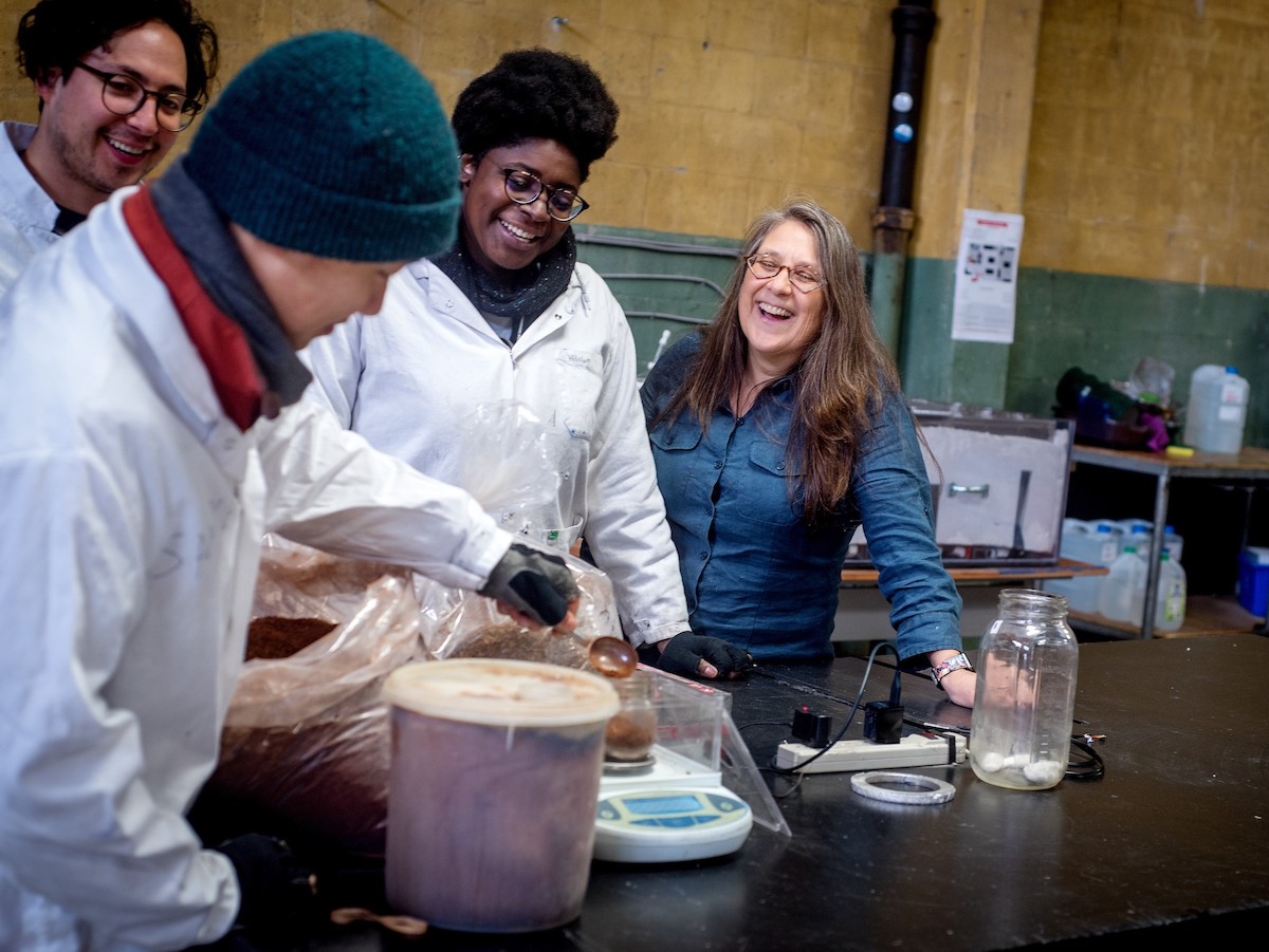 Three people in white coveralls stand over clear plastic bags filled with brown tree bark. Tasha Nathanson looks on, smiling.