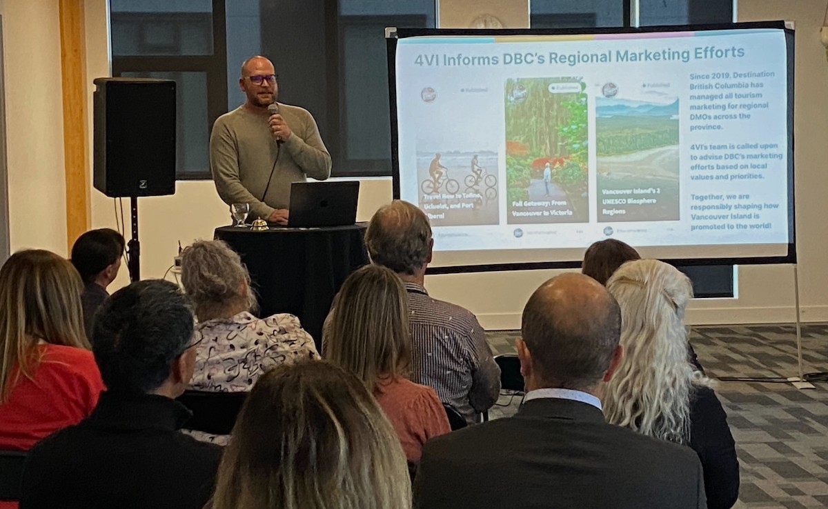 A bald man with a beard and glasses speaks into a microphone in front a group of seated people seen from the back. Beside him a screen shows a slide titled '4VI Informs DBC's Regional Marketing Efforts.'