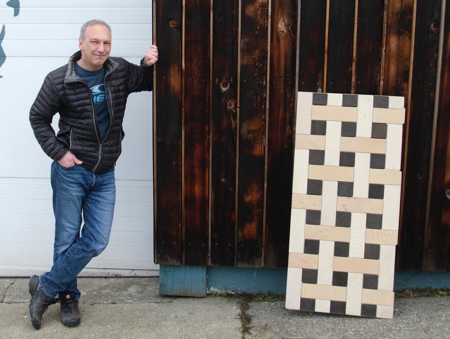 A man with short grey hair and light skin tone, wearing a black puffer jacket and jeans, leans against a wall covered with dark wood boards. A sample of light-coloured wooden tiles arranged in a checker pattern leans against the wall.
